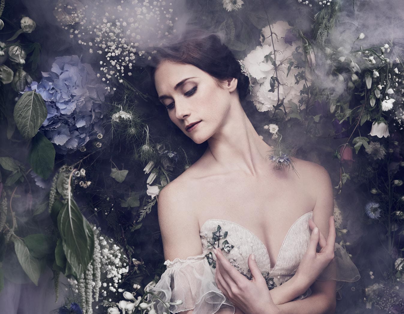 A dancer playing the role of Giselle is shown from the chest up over a Romantic woodland background. She is holding her arms on her chest and looking down while wearing a white dress.