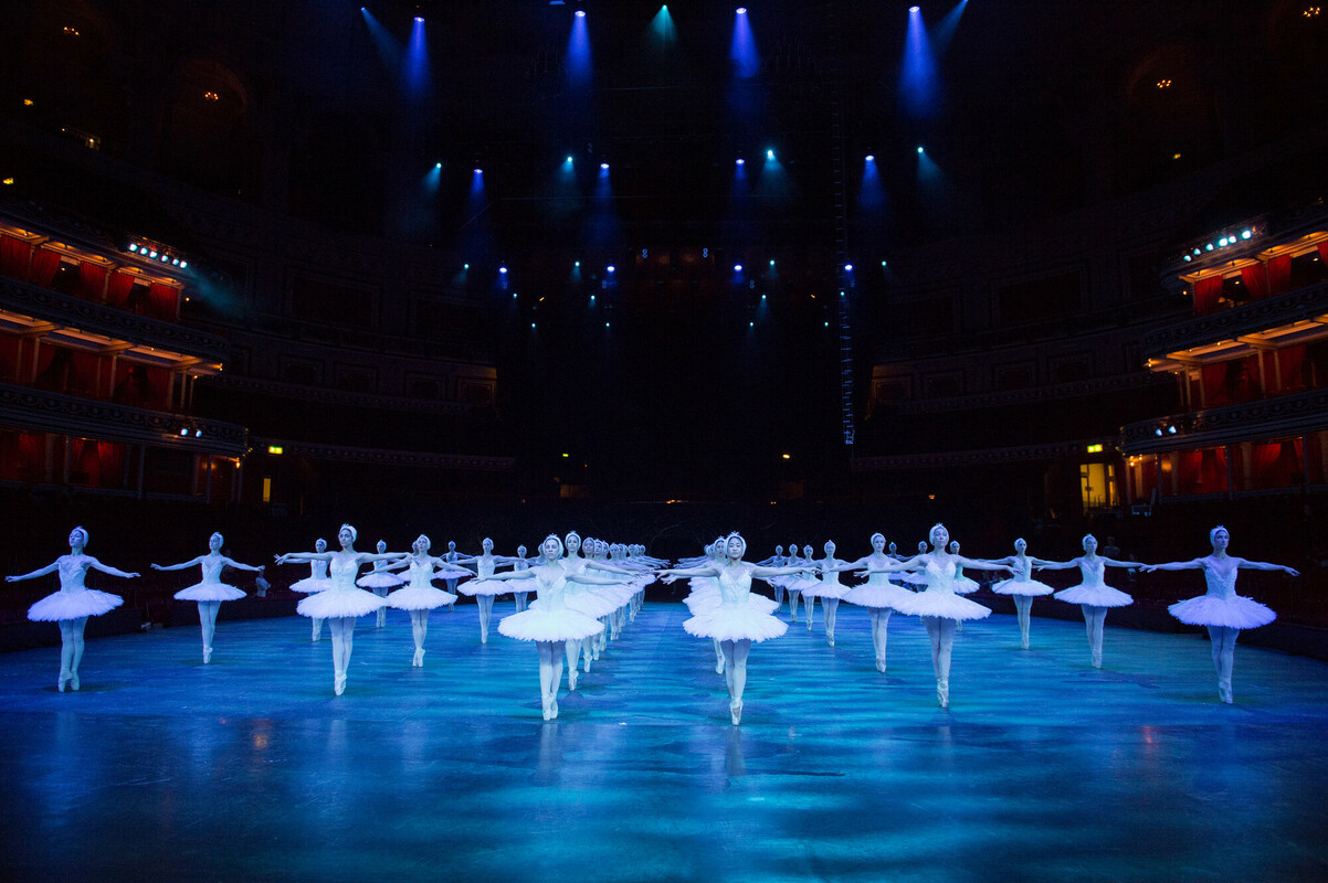 The white swans corps de ballet dancers are show across multiple lines. They are on pointe and wearing white tutus surrounded by atmospheric blue lighting